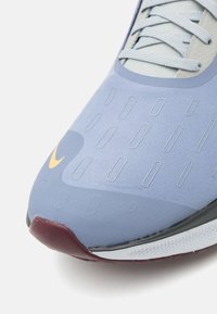 Scarpa sportiva blu con superficie testurizzata, forma aerodinamica, suola più scura in contrasto e logo giallo distintivo.