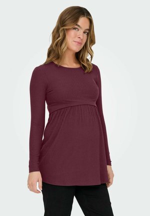 Femme enceinte aux cheveux ondulés portant un haut bordeaux ajusté à manches longues en côtes et un pantalon noir, debout devant un fond uni.