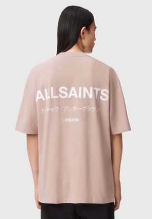 Jasnoróżowa koszulka z krótkim rękawem z dużym białym napisem "ALLSAINTS" i "LONDON" z tyłu. Miękka tkanina o luźnym kroju.