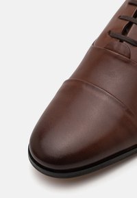 Chaussure en cuir marron avec une texture lisse, bout pointu et détails de couture subtils. Elle est dotée de laçages sombres et d'une semelle noire durable.