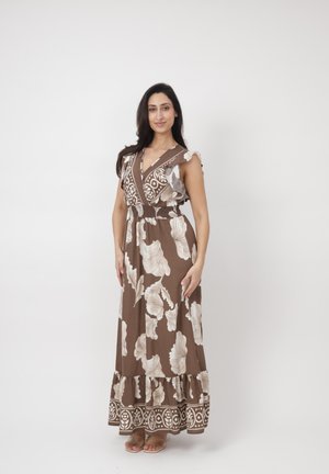 Donna in piedi che sorride, indossa un abito lungo marrone senza maniche con grandi motivi floreali bianchi e sandali.