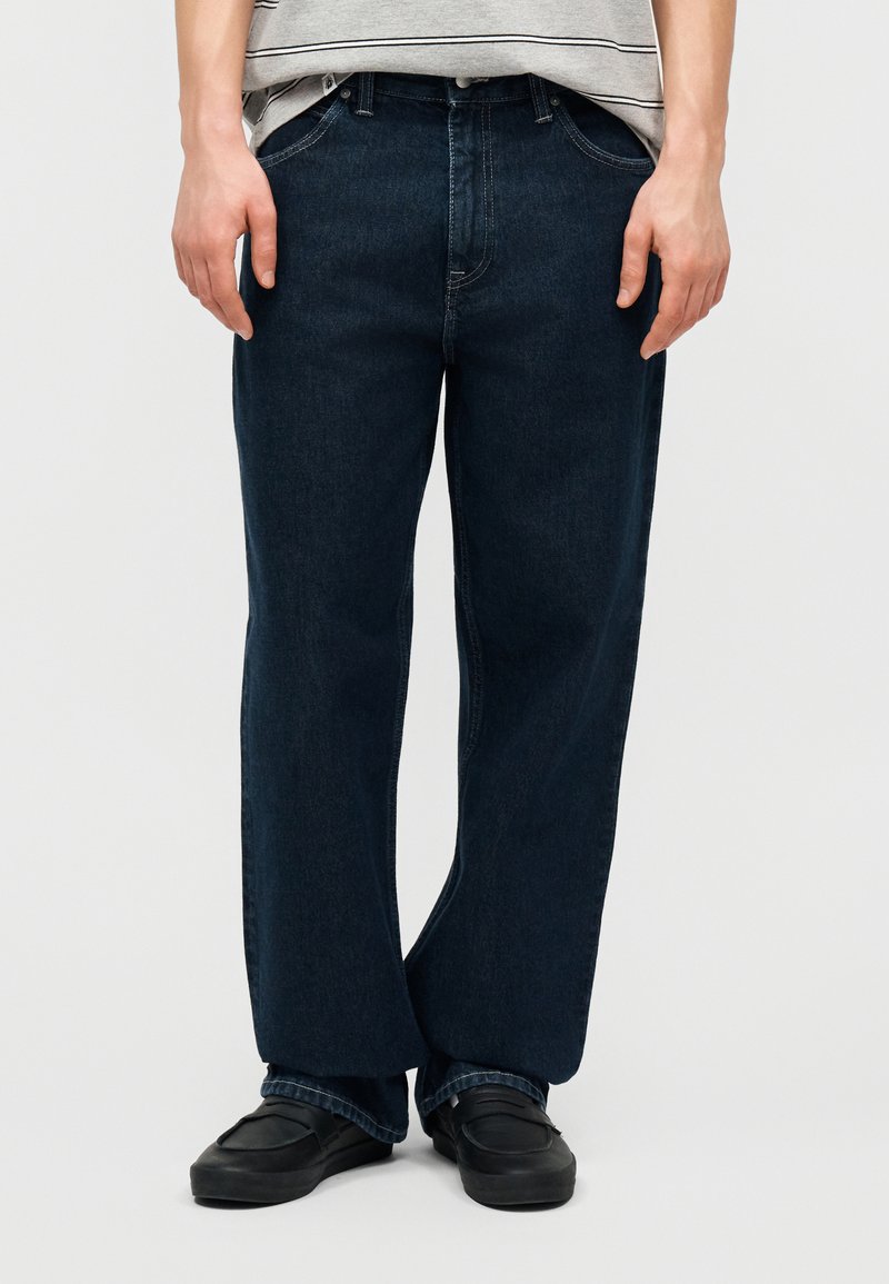 Άτομο που φοράει σκούρα μπλε τζιν με ευθεία γραμμή, μαύρα παπούτσια χωρίς κορδόνια και γκρι ριγέ κοντομάνικη μπλούζα, στέκεται με τα χέρια χαλαρά στα πλάγια.