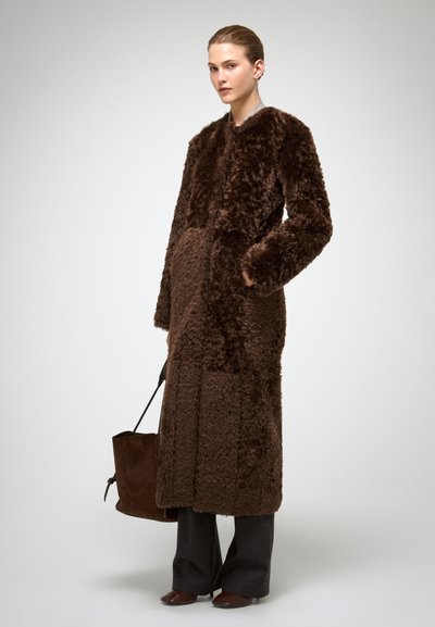 Bruine jas van imitatiebont met lange mouwen, zijzakken en een rechte snit. Draagt over donkergrijze broek, gecombineerd met een bruine handtas.