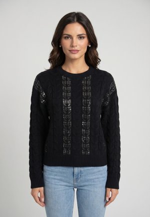 Donna con capelli scuri e ondulati che indossa orecchini di perle, un maglione nero lavorato a maglia con motivi verticali e jeans azzurri, in piedi davanti a uno sfondo semplice.