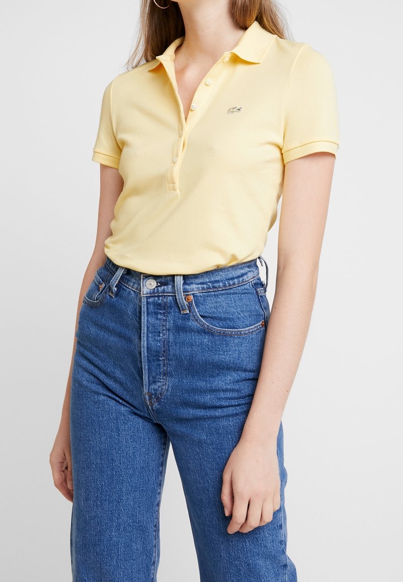 Femme portant un polo jaune pâle à manches courtes rentré dans un jean en denim bleu taille haute sur un fond uni.