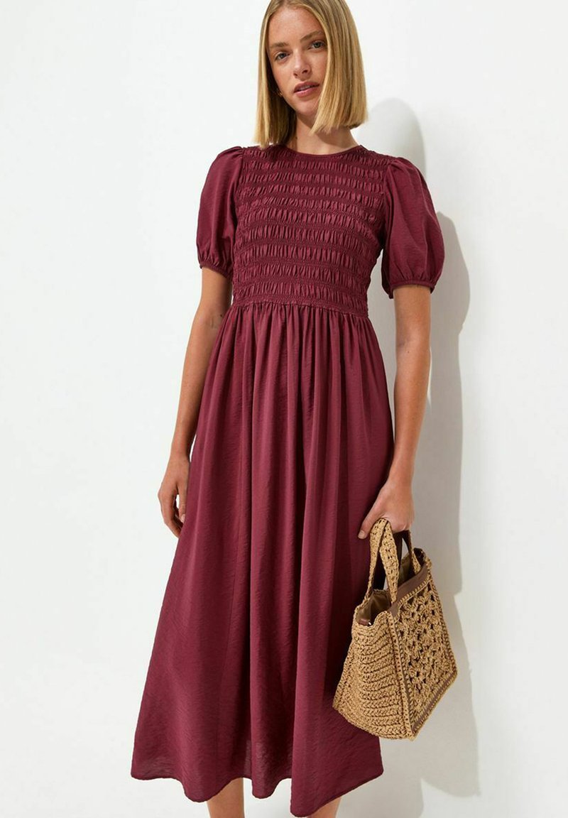Woman wearing a burgundy midi dress with puff sleeves, holding a woven straw handbag, standing against a white wall.