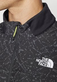 Melns apģērbs ar raibu pelēku rakstu, ar aizsējamu apkakli un neona dzeltenu akcentu. Uz auduma ir balts "The North Face" logo.