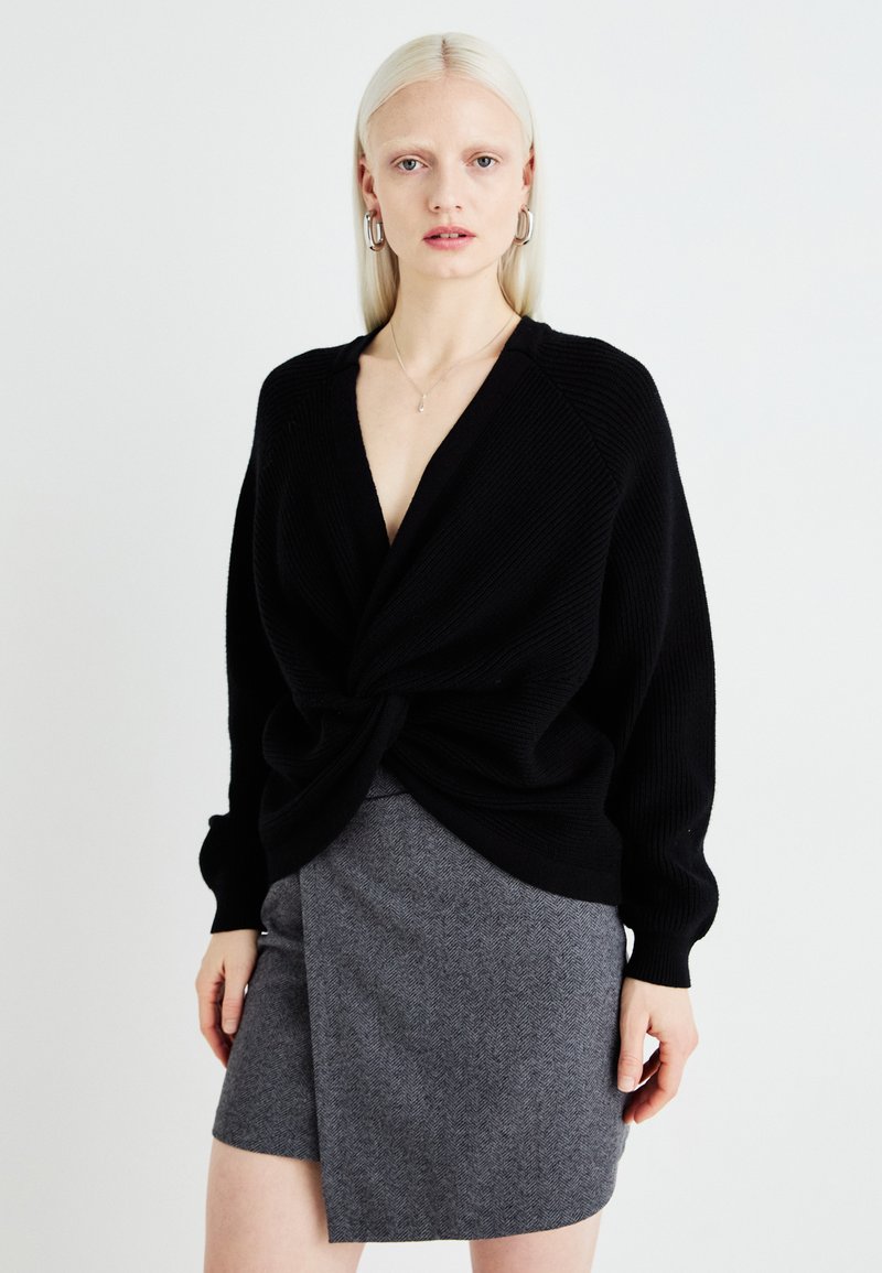 Pulover negru, cu dungi, cu un front răsucit, asortat cu o fustă mini gri cu model herringbone. Are mâneci lungi și un decolteu în V adânc.