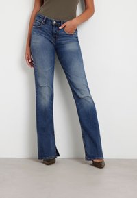 Sinine denimpüksid, millel on laienev lõige, kulunud viimistlus, reite detailid, viie tasku disain ning klassikaline nupuga ja tõmbluku sulgemine.