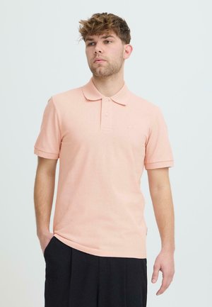 Jeune homme aux cheveux courts, debout, une main dans la poche, vêtu d'un polo rose clair à manches courtes et d'un pantalon noir, sur un fond uni.