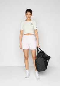 Lys beige avkortet T-skjorte med logo, parret med rosa shorts med sidelommer. Modellen holder en svart treningsbag og har på seg hvite sneakers.