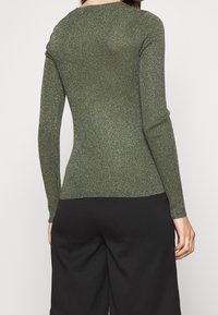 Haut vert côtelé à manches longues avec fils métalliques, coupe ajustée, encolure arrondie, poignets sans couture et un léger éclat, associé à un pantalon noir.