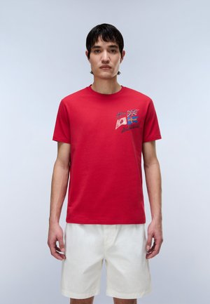 Άνδρας που φοράει κόκκινο μπλουζάκι με τέσσερις μικρές εθνικές σημαίες και λευκό σορτς, στέκεται μπροστά σε απλό φωτεινό φόντο.