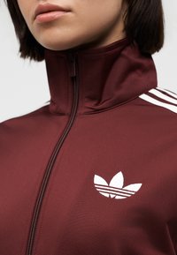 Primer plano de una persona que lleva una chaqueta burdeos con cremallera y cuello alto, con el logo blanco de trébol de Adidas en el pecho.