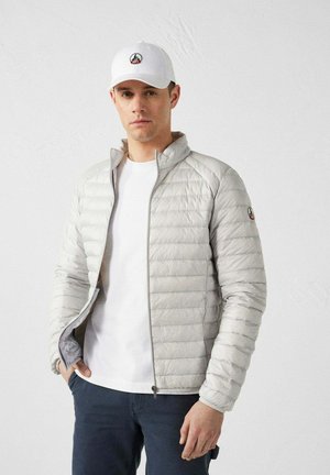 Giacca imbottita grigia chiaro con cuciture orizzontali, colletto alto e zip. Indossata sopra una camicia bianca, completata da un cappellino bianco con logo.