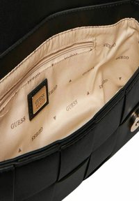 Interior de un bolso negro con un forro beige suave que presenta un patrón repetido de la marca "GUESS" y un bolsillo con cremallera. Exterior texturizado con un diseño tejido.