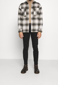 Bikrom plaid flanellipaita valkoisena, mustana ja harmaana, yhdistettynä mustiin slim-fit-housuihin ja ruskeisiin nahkasaappaisiin. Teksturoitu kangas.