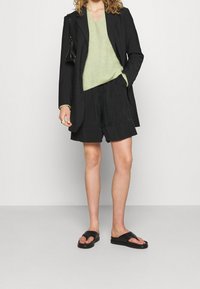 Schwarzer maßgeschneiderter Blazer über einem hellgrünen Strickpullover, kombiniert mit schwarzen Shorts und schwarzen Sandalen. Der Look zeichnet sich durch eine lockere Passform und ein einfaches Design aus.