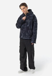 Svart og grå mønstret fleece-hettegenser med glidelås i halsen, sammen med løse svarte cargo-bukser med flere lommer. Hvit sneakers.