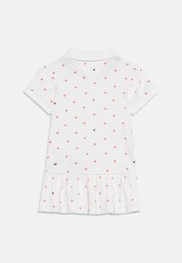 Λευκό φόρεμα με κοντά μανίκια, μικρό κόκκινο σχέδιο καρδιάς και βολάν στο τελείωμα, με κολάρο πόλο, φαίνεται από πίσω σε λευκό φόντο.