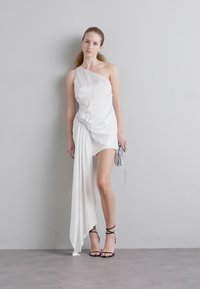 Wit één-schouder jurk met gedrapeerde stof en plooien, met een zilveren embellissement op de taille, gecombineerd met een metallic clutch.