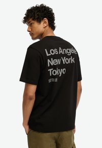 Joven con cabello rizado que lleva una camiseta negra con "Los Ángeles, Nueva York, Tokio" y caracteres japoneses en la espalda, con las manos en los bolsillos.