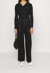 Czarny długi rękaw V-neck top z logo Lacoste, w połączeniu z czarnymi spodniami o wysokim stanie z szerokimi nogawkami i białym przeszyciem oraz teksturowaną czarną torbą.