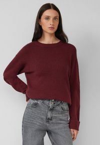 Vrouw met lang zwart haar, gekleed in een bordeauxrode trui en hoogsluitende grijze jeans, staande tegen een effen lichte achtergrond.
