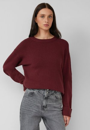 Woman with long dark hair wearing a burgundy sweater and high-waisted grey jeans, standing against a plain light background.