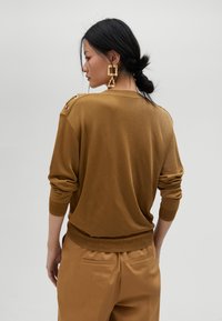 Maglione marrone lavorato a maglia con maniche arrotolate e bottoni sulle spalle, abbinato a pantaloni larghi coordinati. Texture liscia, vestibilità casual.