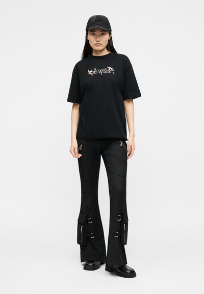 Νεαρή γυναίκα που φοράει μαύρο καπέλο, μαύρο μπλουζάκι Off-White™ με σχέδιο πεταλούδας, μαύρο παντελόνι τύπου cargo με φερμουάρ και αγκράφες και μαύρα παπούτσια.