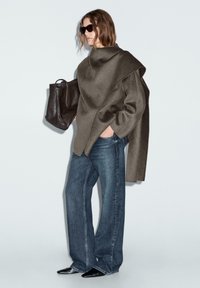 Oversized olijfgroene top met asymmetrisch ontwerp, gecombineerd met wijduitlopende blauwe denim jeans. Een zwarte tote bag en spitse zwarte schoenen maken de look compleet.