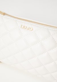 Borsa a tracolla bianca trapuntata con hardware dorato, chiusura a zip e logo del marchio impresso. Presenta una texture liscia e lucida e un motivo geometrico.