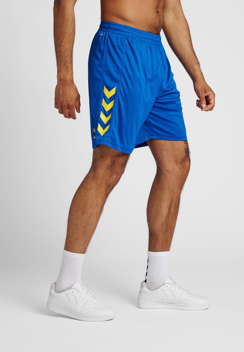 Ein oberkörperfreier Mann, der blaue Sportshorts mit gelben Chevronen, weiße Socken und weiße Sneaker trägt, steht vor einem schlichten Hintergrund.