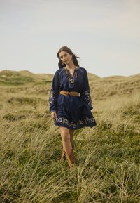 Abito blu navy con maniche lunghe e dettagli floreali ricamati, stretto in vita da una cintura beige, abbinato a stivali alla caviglia marroni.