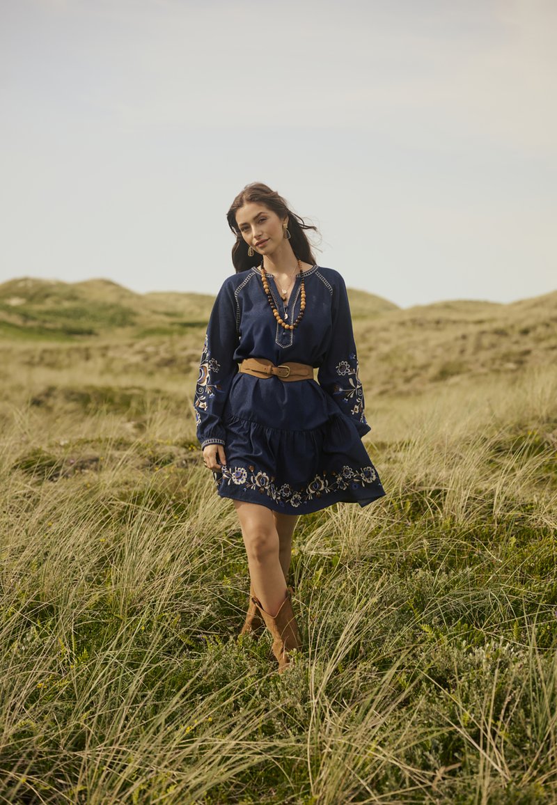 Abito blu navy con maniche lunghe e dettagli floreali ricamati, stretto in vita da una cintura beige, abbinato a stivali alla caviglia marroni.