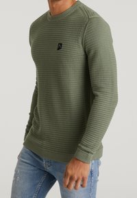 Sweter w oliwkowej zieleni z fakturą wafelka, okrągły dekolt oraz długie rękawy. Na piersi znajduje się małe czarne logo.