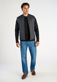 Svart stickad långärmad tröja, grå vadderad väst med vertikal söm, ljusblå rak ben jeans, bruna skor med snörning.