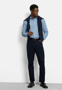 Lichtblauw overhemden met knopen, donkerblauwe broek, een donkere trui over de schouders gedrapeerd, zwarte schoenen, staande pose, minimalistische achtergrond.