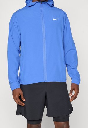 Homme portant une veste à capuche bleue Nike avec fermeture éclair et un short de sport noir par-dessus un short de compression, debout devant un fond blanc.