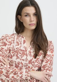 Jeune femme aux longs cheveux bruns, bras croisés, portant un chemisier rose clair avec un motif floral rouge, se tenant devant un arrière-plan uni clair.