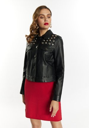 Frau trägt eine schwarze Lederjacke mit Metallnieten auf den Schultern und einen roten Rock, steht vor einem schlichten hellen Hintergrund.