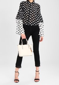 Woman wearing black and white polka dot blouse with tiered sleeves, black cropped fringe pants, black heels, holding a white tote bag.