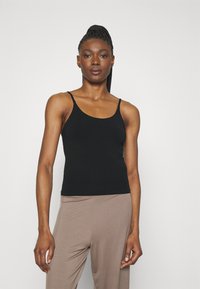 Black tank top with thin straps, fitted design, made from a smooth fabric. Paired with light brown, loose-fitting pants.