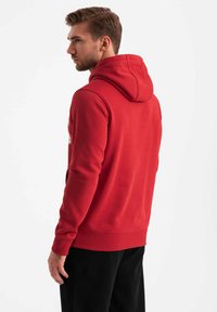 Röd hoodie i fleece-material, med en dragsko i huvan, ribbade muddar och en framficka. Tjock textur med en enkel design.