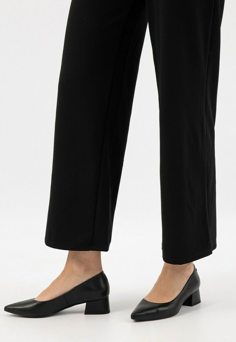 Escarpins en cuir noir avec un bout pointu et un talon block, présentant une texture lisse et un design minimaliste, associés à un pantalon noir.