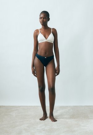 Modella con pelle scura e capelli intrecciati in piedi a piedi nudi, indossa un bralette bianco Tommy Hilfiger e mutandine blu navy su uno sfondo semplice.