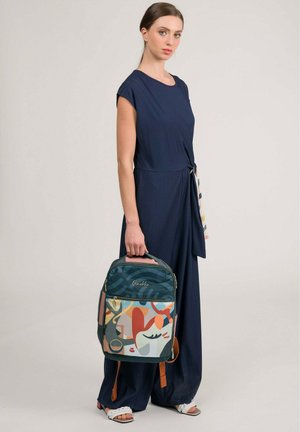 Femme en combinaison bleu marine tenant un sac à dos coloré à motif par sa poignée, debout devant un fond clair uni.