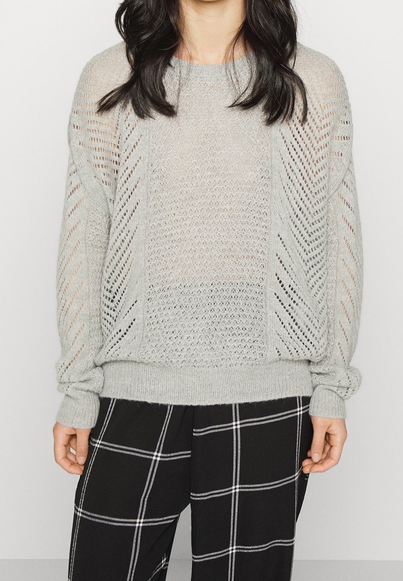 Femme portant un pull en tricot gris clair avec des motifs en chevrons ajourés et un pantalon large noir à carreaux blancs en damier.