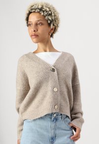 Beige gestrickte Strickjacke mit V-Ausschnitt, drei dekorativen Knöpfen, geripptem Saum und Bündchen sowie weicher, flauschiger Textur. Tragen über einem weißen Hemd.